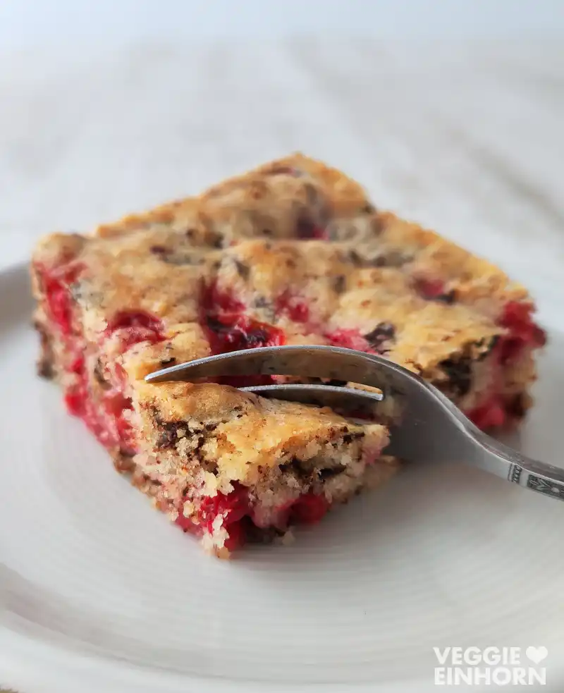 Ein Stück veganer Nusskuchen mit Johannisbeeren und Schokolade auf einem Teller, eine Gabel steckt im Kuchen.