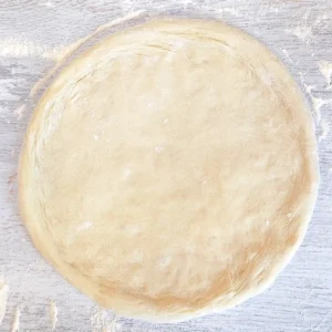 Ein veganer runder Pizzateig mit einem Rand liegt fertig vorbereitet auf einer bemehlten Arbeitsfläche.