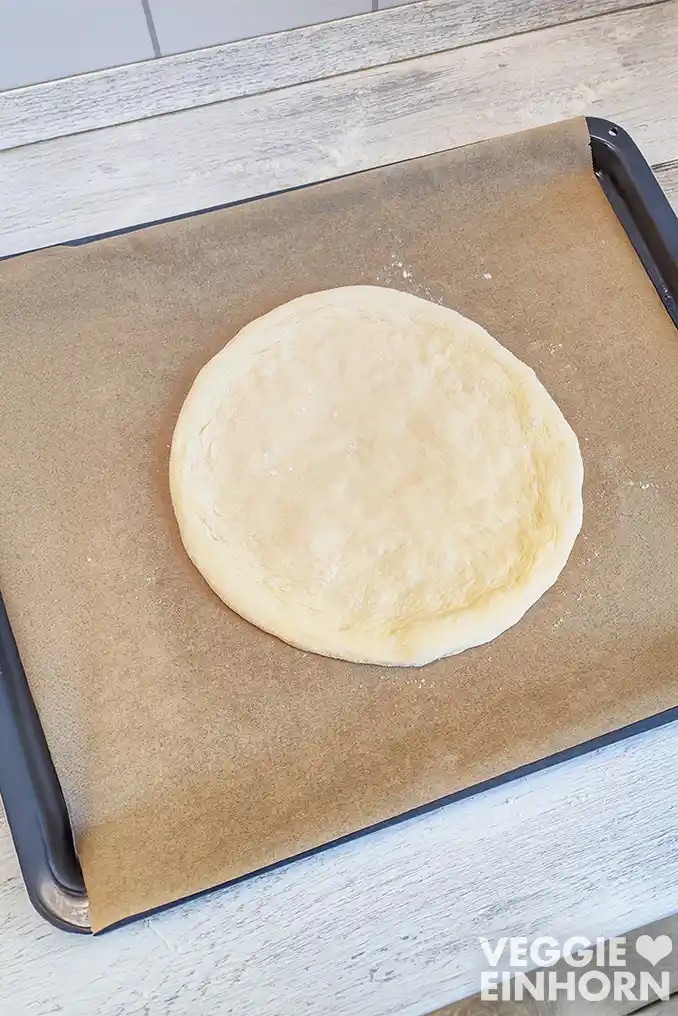 Ein runder veganer Pizzateig mit einem Rand liegt auf einem mit Backpapier ausgelegten Backblech.