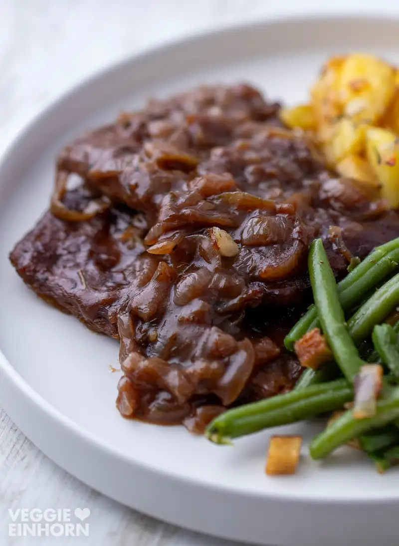 Nahaufnahme von veganem Rostbraten aus Soja Big Steaks mit Zwiebelsauce, Bratkartoffeln und grünen Bohnen auf einem weißen Teller.