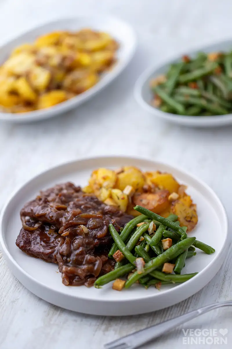 Weißer Teller mit veganem Rostbraten aus Soja Big Steaks, Bratkartoffeln und grünen Bohnen, im Hintergrund Schüsseln mit Beilagen.