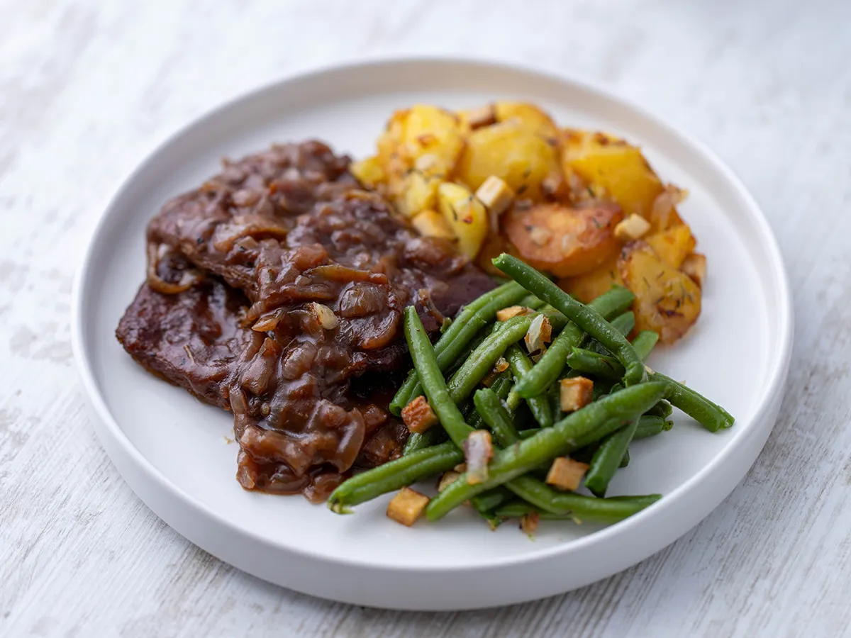 Veganer Rostbraten aus Soja Big Steaks mit Zwiebelsoße auf einem Teller mit Bratkartoffeln und grünen Bohnen.