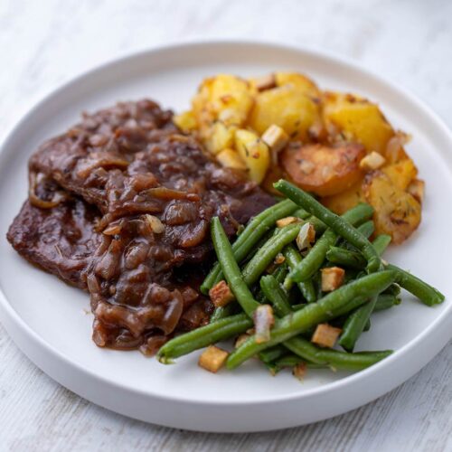 Nahaufnahme von veganem Rostbraten aus Soja Big Steaks mit Zwiebelsauce, Bratkartoffeln und grünen Bohnen.