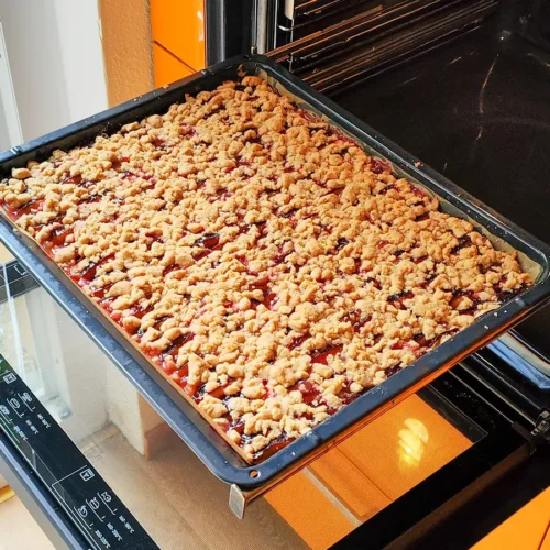 Ein großes Backblech mit veganem Zwetschgenkuchen, der mit goldbraunen Zimtstreuseln bedeckt ist und im geöffneten Backofen steht.