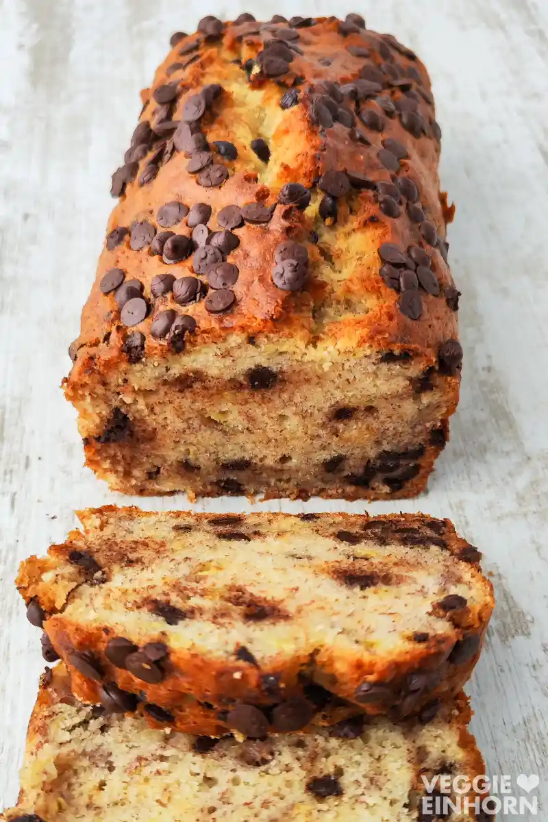 Veganes Bananenbrot mit Schokolade Ein veganes Bananenbrot mit vielen Schokotropfen liegt angeschnitten auf einem hellen Holztisch.