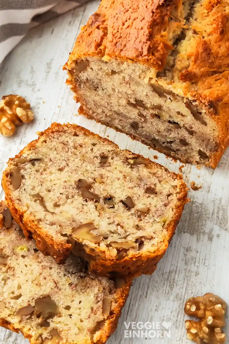 Veganes Bananenbrot mit Walnüssen Ein angeschnittenes veganes Bananenbrot mit Walnüssen liegt auf einer hellen Holzoberfläche, daneben liegen einzelne Walnusskerne.