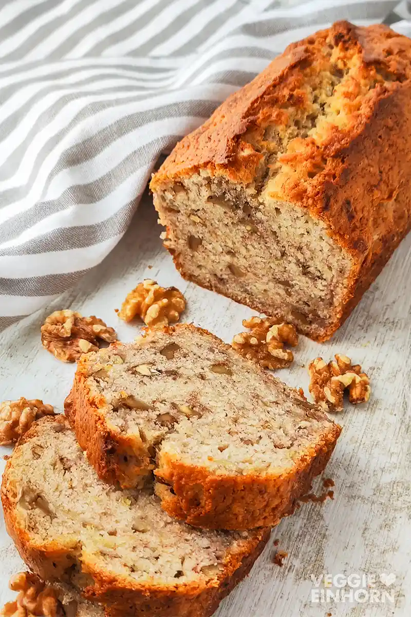 Ein angeschnittenes veganes Bananenbrot mit Walnüssen liegt auf einem hellen Tisch neben einem gestreiften Tuch und einzelnen Walnüssen.
