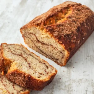 Ein angeschnittenes veganes Bananenbrot mit Zimt auf einem hellen Holztisch.