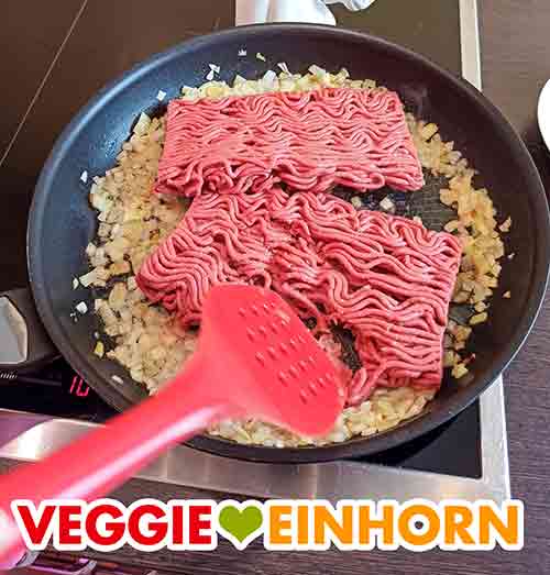 Veganes Hackfleisch in der Pfanne mit den Zwiebeln Das vegane Hack in der Pfanne wird mit einem Pfannenwender zerteilt.