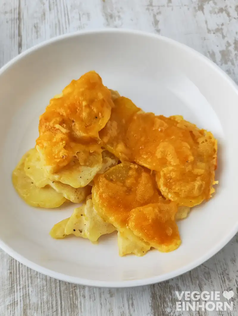 Eine Portion veganes Kartoffelgratin mit veganem Käse liegt auf einem weißen Teller auf hellem Holztisch.