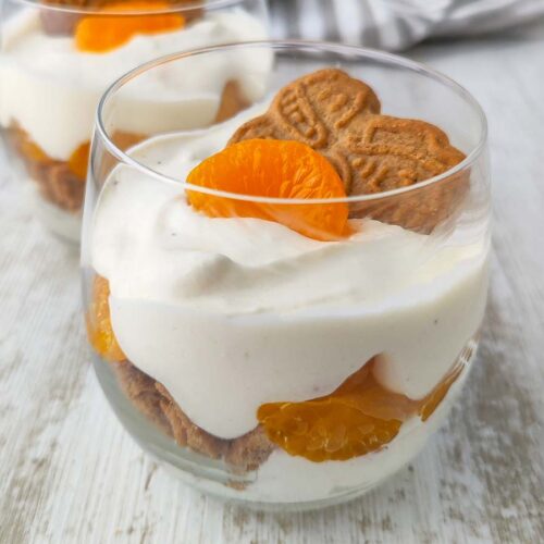 Ein veganes Schichtdessert im Glas mit Dessertcreme, Mandarinen und Spekulatius, fotografiert auf hellem Untergrund.