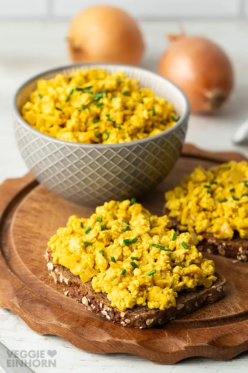 Brot mit veganem Rührei, im Hintergrund eine Schale mit veganem Rührei und Zwiebeln.