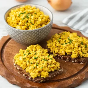 Zwei Brote mit veganem Rührei und eine Schale mit veganem Rührei auf einem runden Holzbrett.