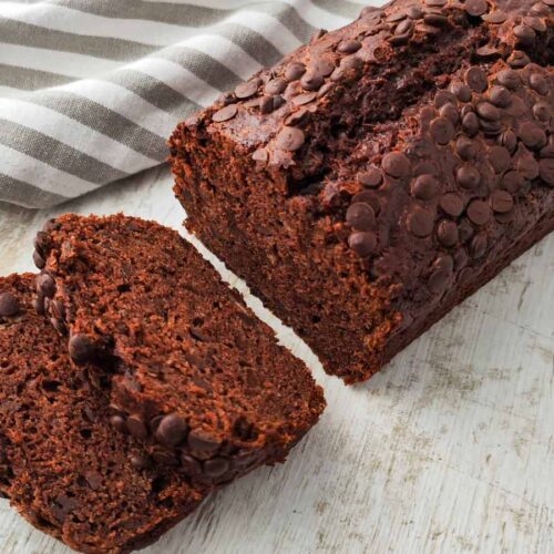 Ein veganes Schoko-Bananenbrot liegt mit zwei abgeschnittenen Scheiben auf einer hellen Fläche, obenauf befinden sich viele Schokotropfen.