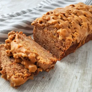 Angeschnittenes veganes Spekulatius Bananenbrot mit Karamellglasur auf einem hellen Holztisch, daneben ein gestreiftes Tuch.