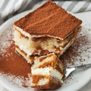 Ein Stück veganes Tiramisu mit cremiger Füllung und getränktem Biskuit ist mit Kakaopulver bestäubt und auf einem weißen Teller angerichtet.