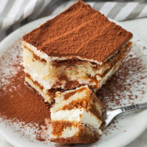 Ein Stück veganes Tiramisu mit dicker Kakaoschicht auf der Oberfläche liegt auf einem weißen Teller.