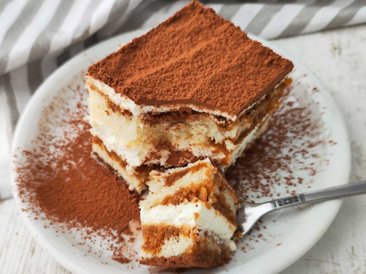 Ein Stück veganes Tiramisu mit dicker Kakaoschicht auf der Oberfläche liegt auf einem weißen Teller.