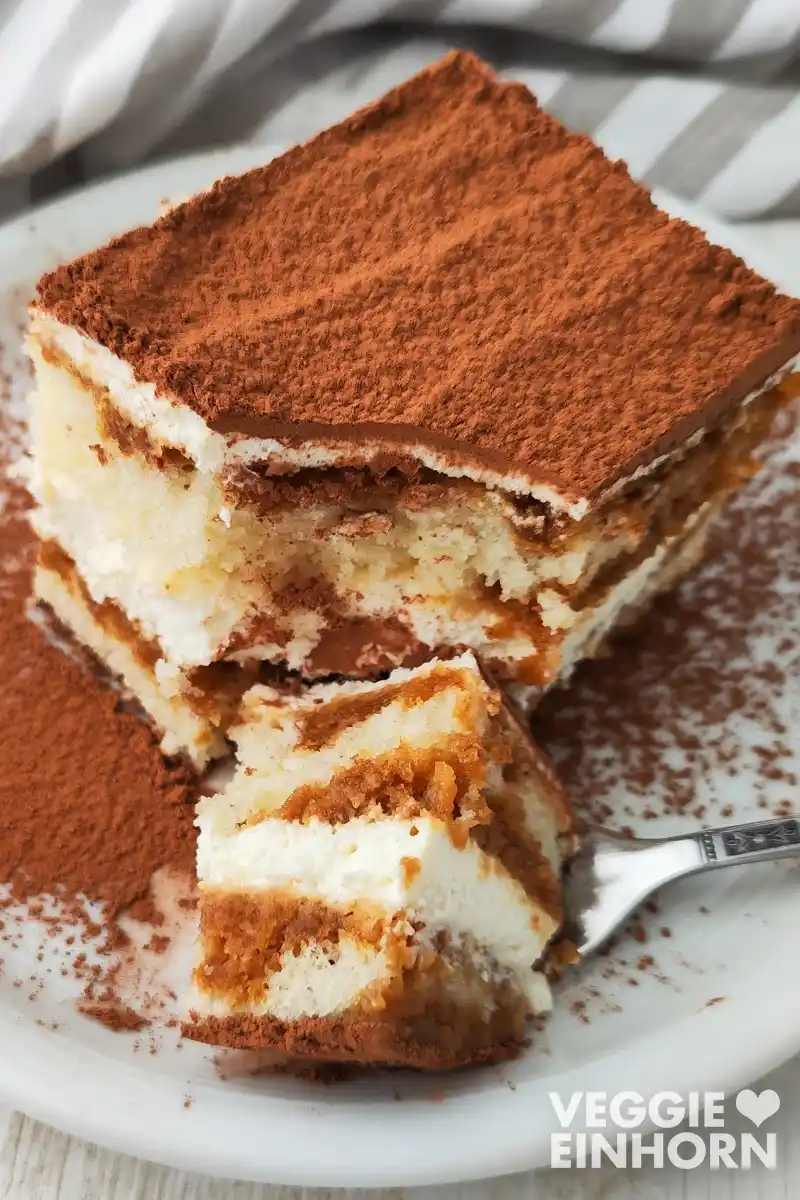 Ein Stück veganes Tiramisu liegt auf einem weißen Teller, im Vordergrund ein Stück des Tiramisus auf einer Gabel.
