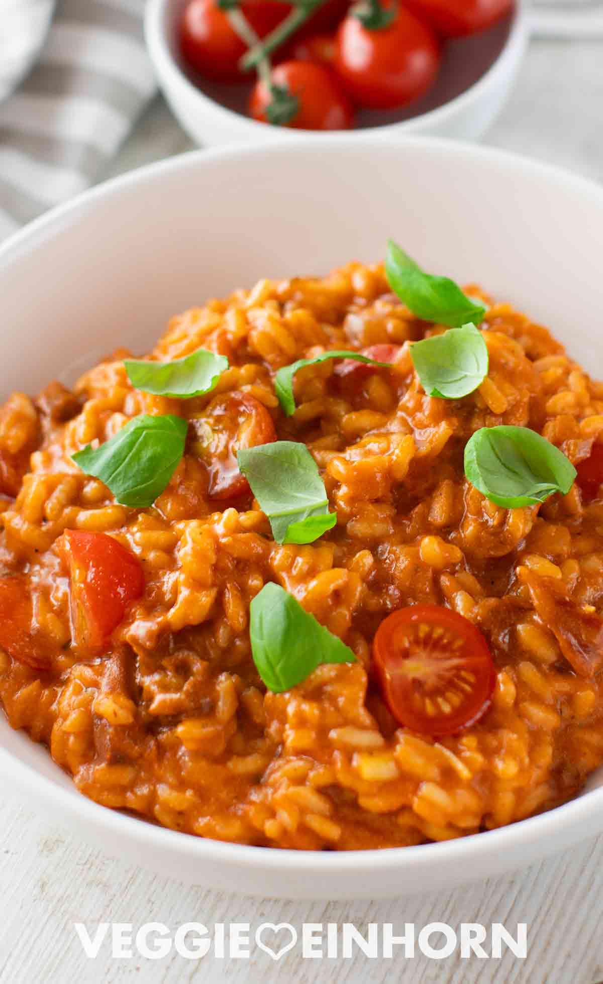 Veganes Tomatenrisotto in einer weißen Schale, garniert mit frischem Basilikum und Kirschtomaten, im Hintergrund eine Schale mit frischen Tomaten.