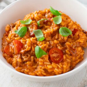 Veganes Tomatenrisotto, garniert mit frischem Basilikum und halbierten Kirschtomaten in einer weißen Schale.