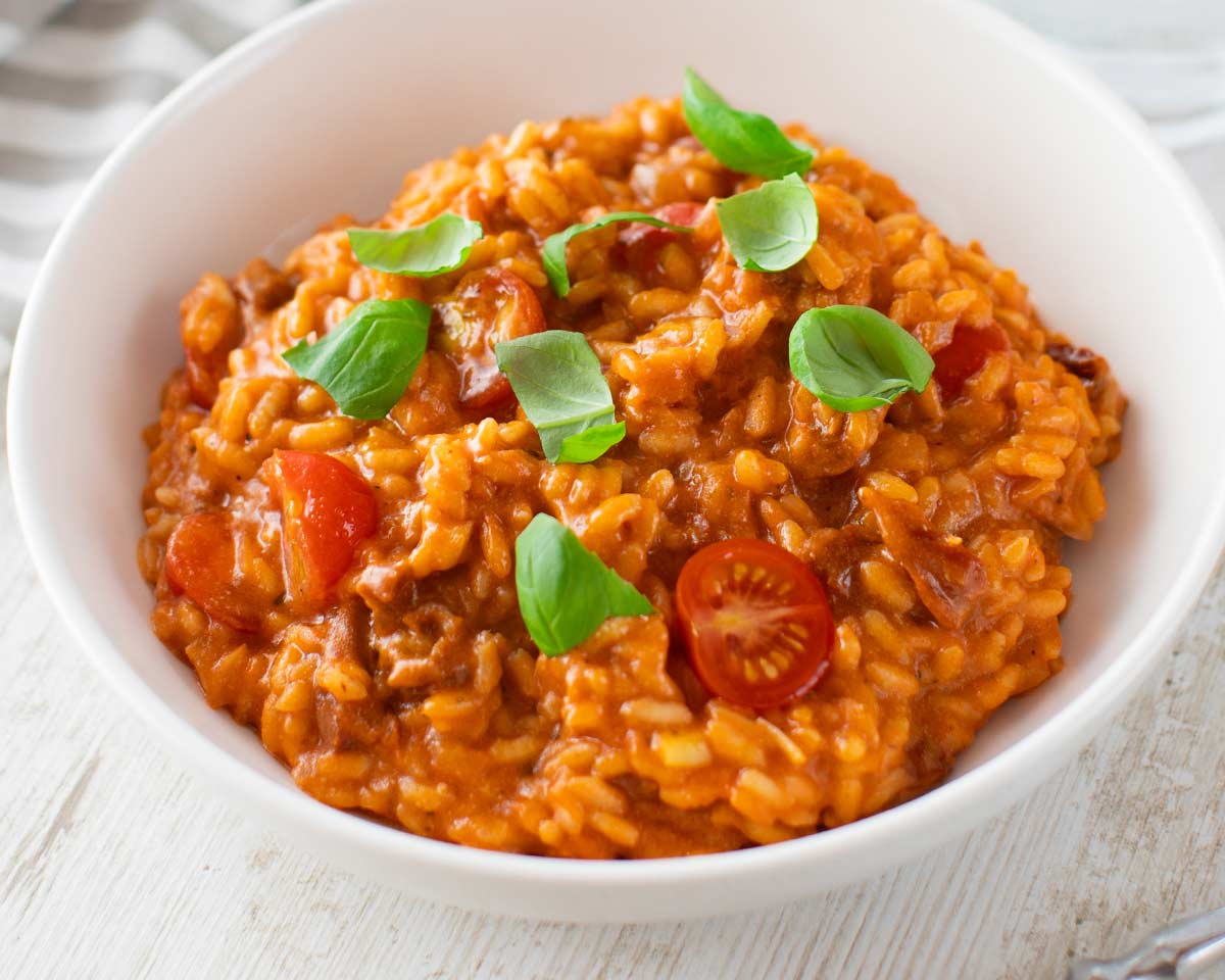Veganes Tomatenrisotto, garniert mit frischem Basilikum und halbierten Kirschtomaten in einer weißen Schale.