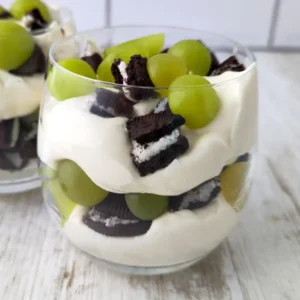 Zwei Gläser mit veganem Nachtisch aus Dessertcreme, grünen Weintrauben und zerbrochenen Oreo Keksen auf einem hellen Holztisch.