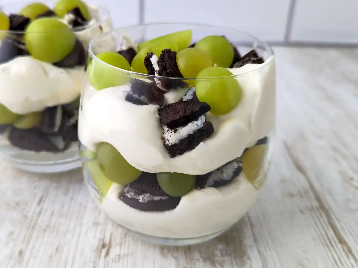 Zwei Gläser mit veganem Nachtisch aus Dessertcreme, grünen Weintrauben und zerbrochenen Oreo Keksen auf einem hellen Holztisch.