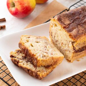 Ein veganer Zimtkuchen mit Apfel, angeschnitten auf einer weißen Kuchenplatte. Im Hintergrund liegen frische Äpfel und Zimtstangen.