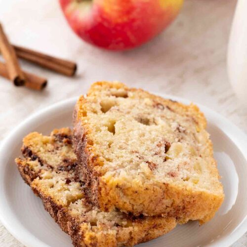 Nahaufnahme von zwei Scheiben veganem Zimtkuchen mit Apfelstücken, serviert auf einem weißen Teller. Im Hintergrund sind frische Äpfel und Zimtstangen zu sehen.