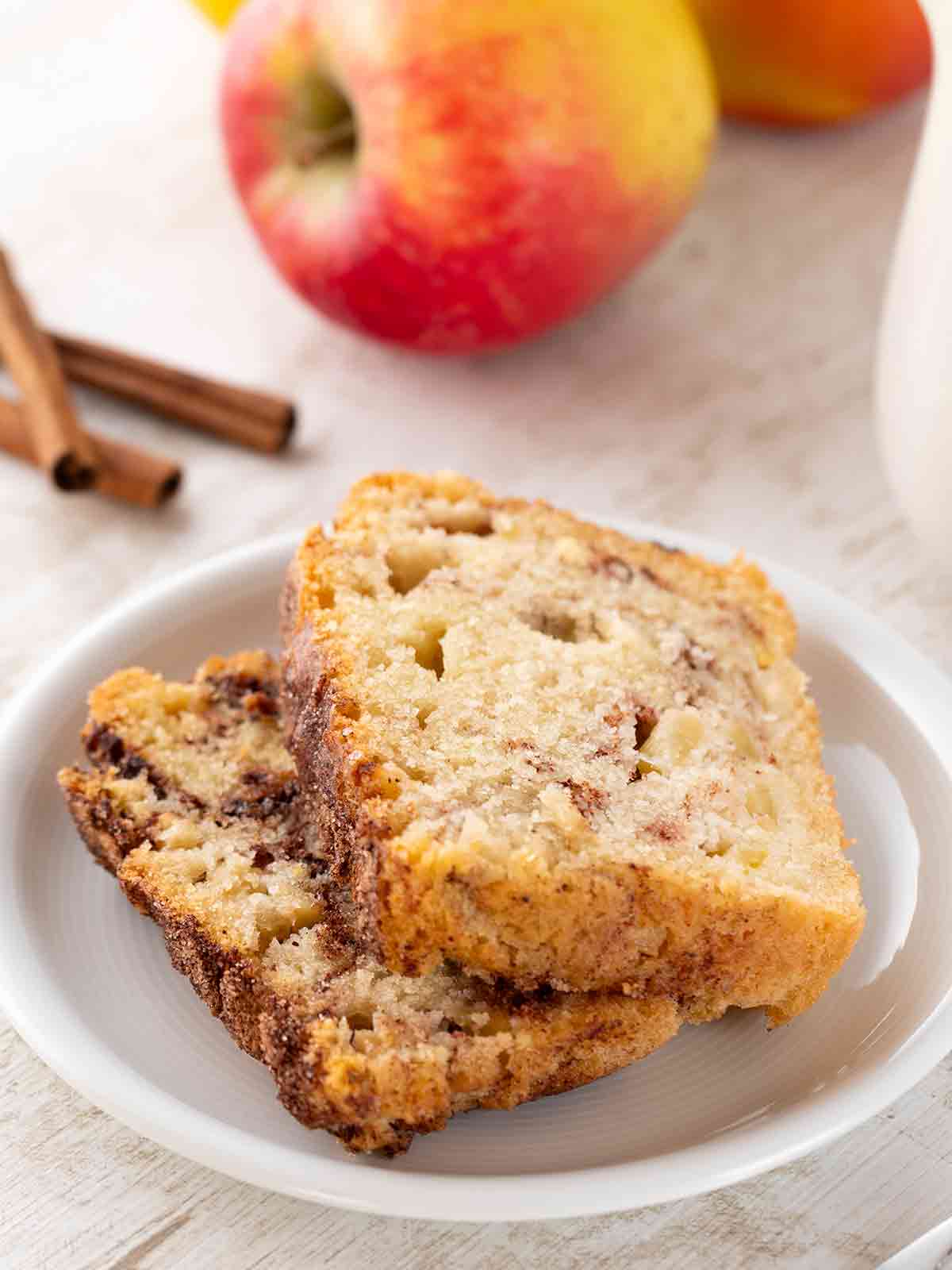 Nahaufnahme von zwei Scheiben veganem Zimtkuchen mit Apfelstücken, serviert auf einem weißen Teller. Im Hintergrund sind frische Äpfel und Zimtstangen zu sehen.