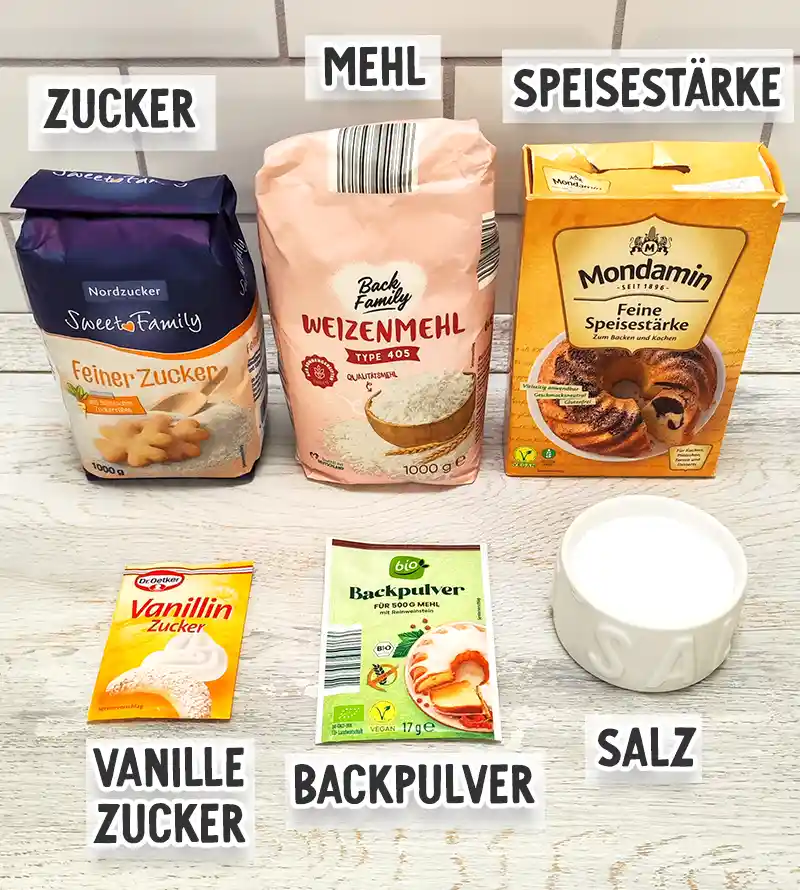 Verschiedene Backzutaten wie Zucker, Weizenmehl, Speisestärke, Vanillezucker, Backpulver und Salz stehen beschriftet auf einer hellen Arbeitsfläche.